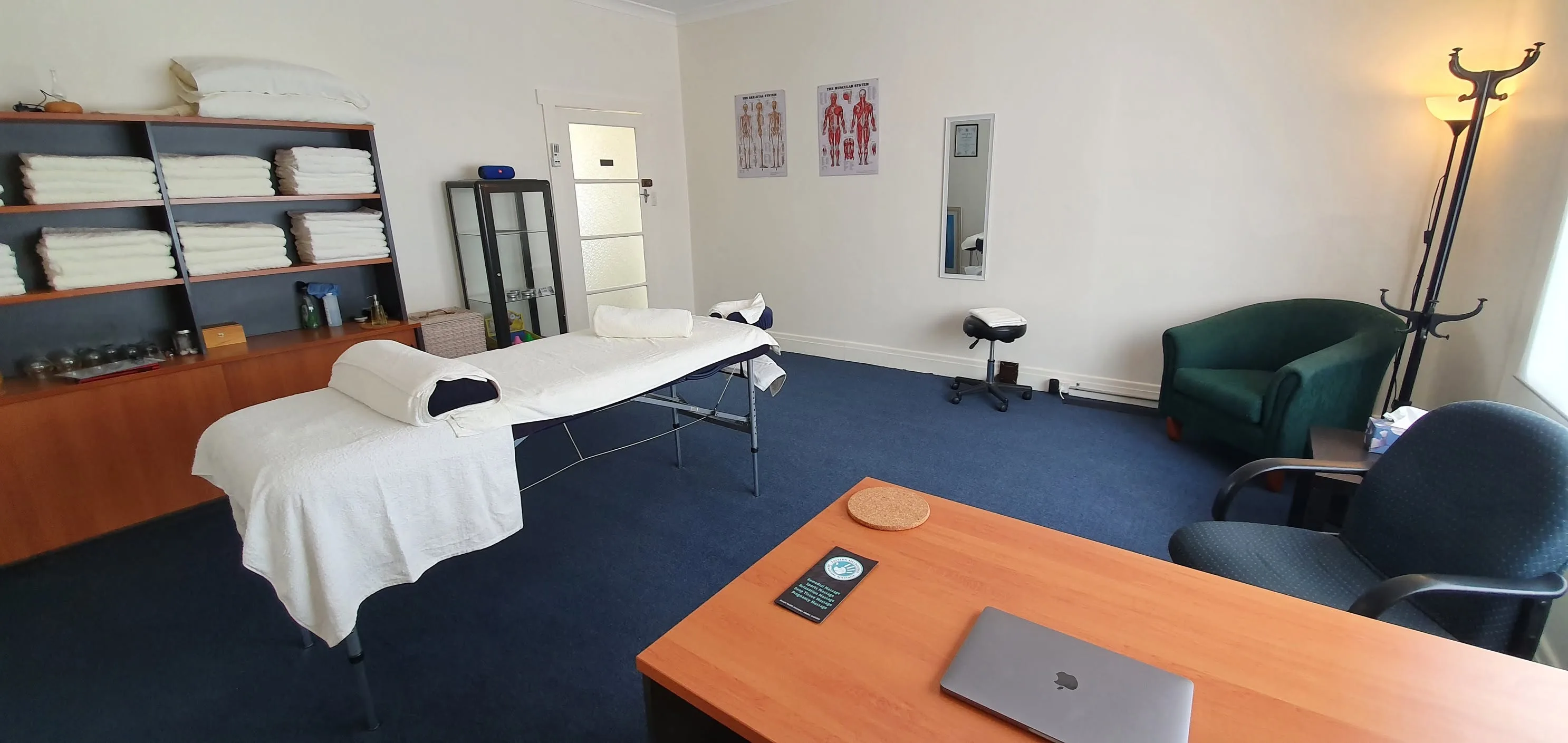 The treatment room — made-up massage table, shelves of rolled white towels, anatomy posters and a green reading chair