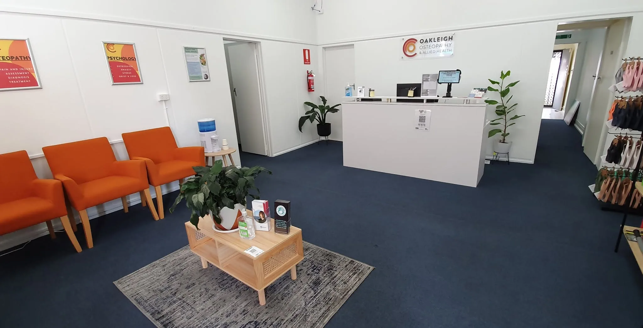 Shared reception and waiting area — orange tub chairs, white reception desk under the Oakleigh Osteopathy wall sign, blue carpet and potted plants