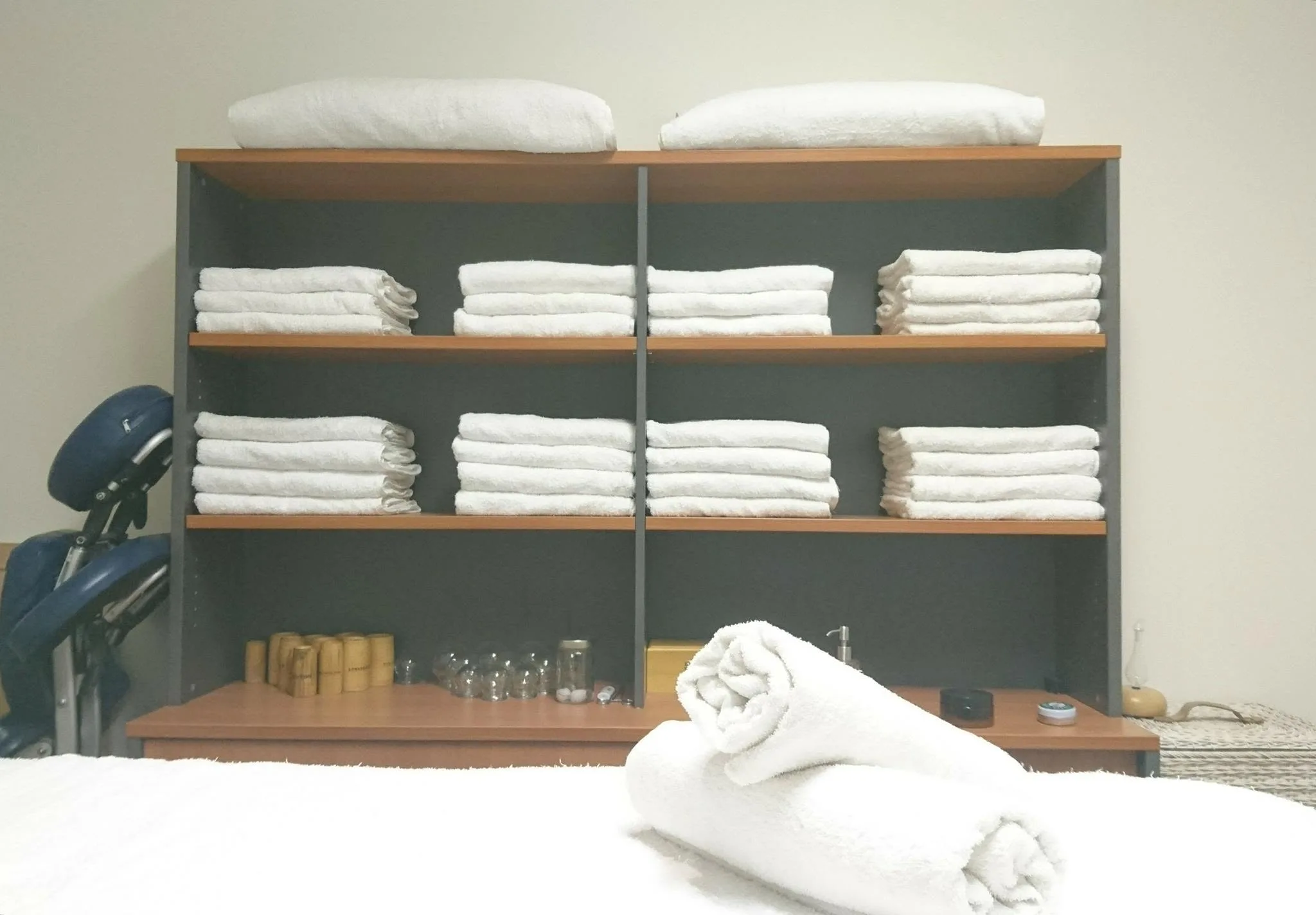 Clinic detail — neat shelves of rolled white towels, cupping cups and product tins, and a portable massage chair
